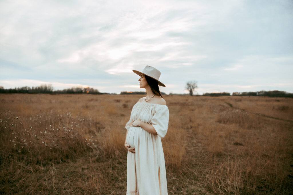 Pregnant woman holding her baby bump in a cream colored dress against the brown landscape in the midwest.